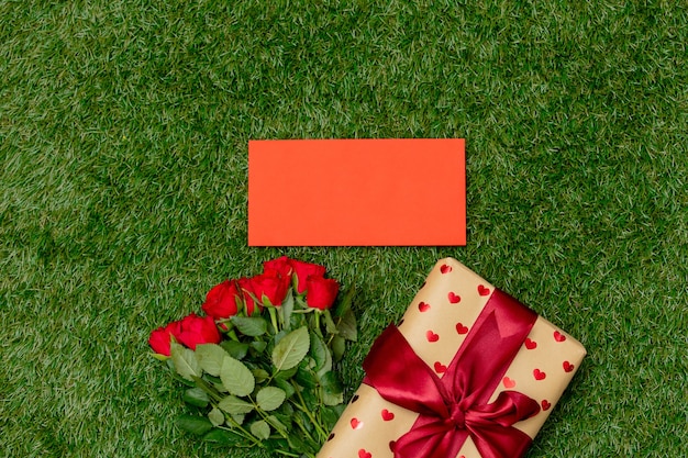 Red roses bouquet and gift box with envelope on green grass.