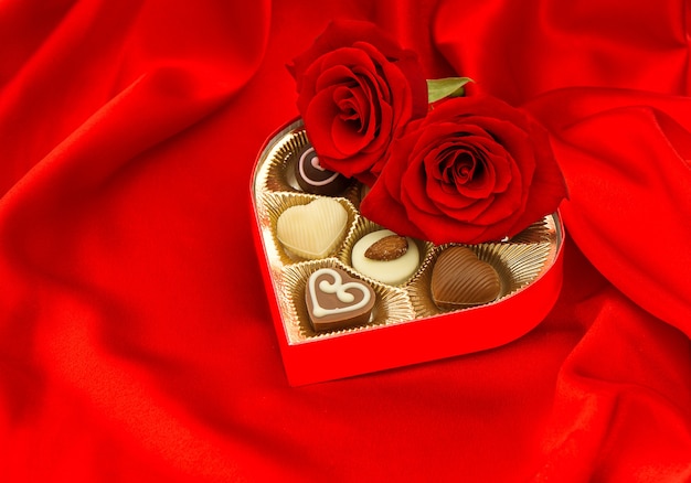 Rose rosse e praline di cioccolato in confezione regalo dorata a forma di cuore su fondo satinato