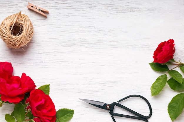 Red roses, jute rope and scissors on white wooden table, top view