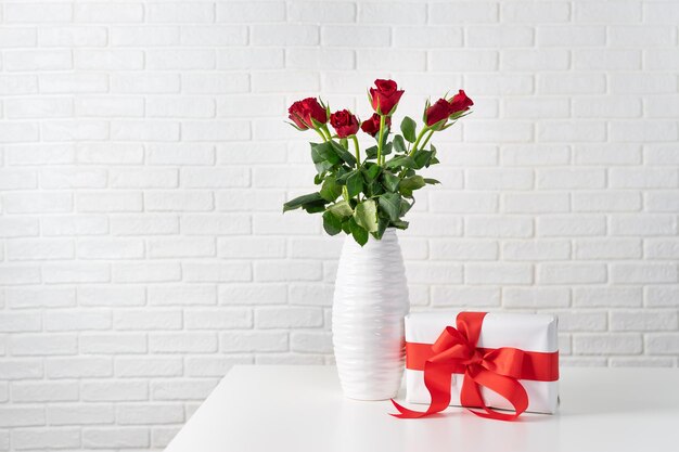 Photo red roses in white vase next to a gift box with red ribbon against white brocks wall