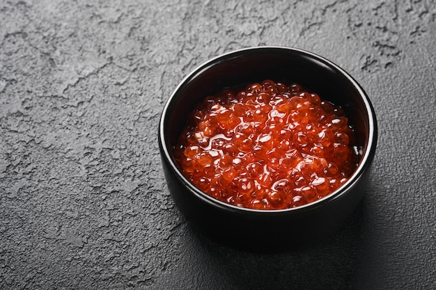 Foto caviale di salmone rosso delizioso caviale rosso in ciotola nera su sfondo nero in cemento scuro vista dall'alto con spazio per la copia posa piatta mock up