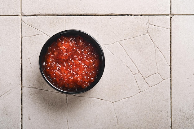 Red salmon caviar Delicious red caviar in black bowl on old cracked tile table background Top view with copy space Flat lay Mock up