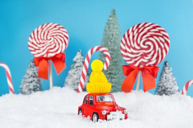 Automobile rossa della santa con il berretto invernale giallo e la foresta dell'albero di natale con caramelle e lecca-lecca
