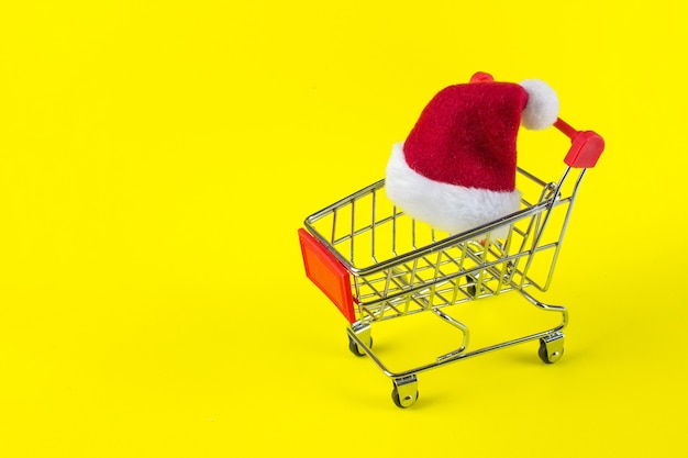 Red Santa's Christmas hat on yellow background in trolley cart on yellow background.