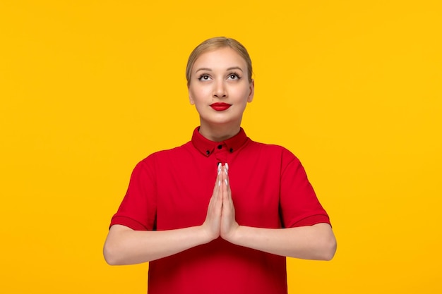 Ragazza carina giorno della camicia rossa che si tiene per mano e prega in una camicia rossa su sfondo giallo