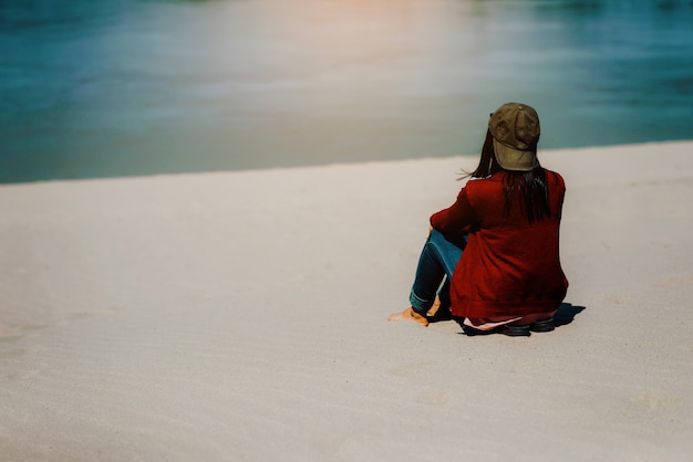 A red shirt woman sitting on a sandy beach facing the river or the sea.