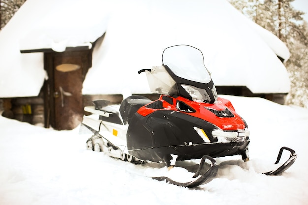 Red snowmobile is standing on a snow on the background of a winter house log hut and forest