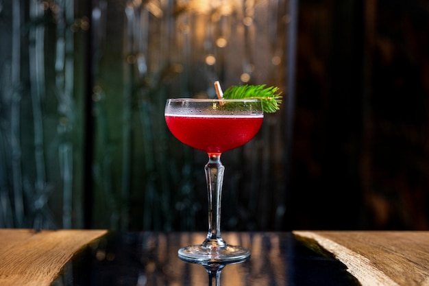 A red sour cocktail in a coupe glass, decorated with a fir branch on the table in a bar