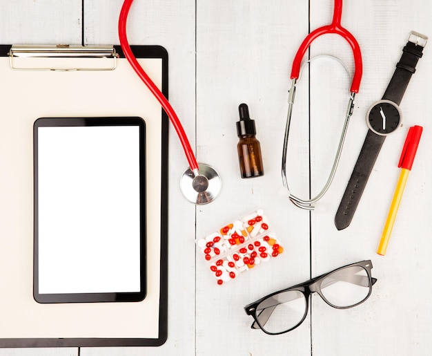 Red stethoscope clipboard smartphone glasses and pills on white wooden table