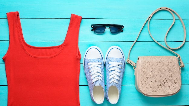 Red T-shirt, sneakers, leather bag, sunglasses on a blue wooden table.