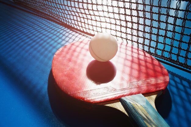 Una racchetta da ping pong rossa e una palla bianca giacciono sulla superficie del tavolo accanto alla rete sport gioco