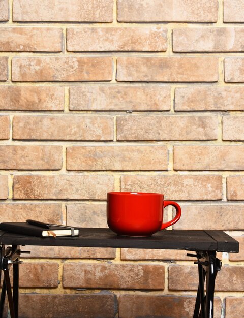 Red tea or coffee cup or ceramic mug for drink copybook and pen black tray on brick textured background copy space