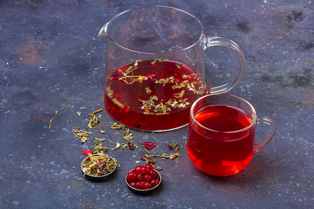 Red tea (rooibos, hibiscus, karkade) in glass cup and teapot among dry tea leaf, petals and cranberries on a dark background. Herbal, vitamin, detox tea for cold and flu