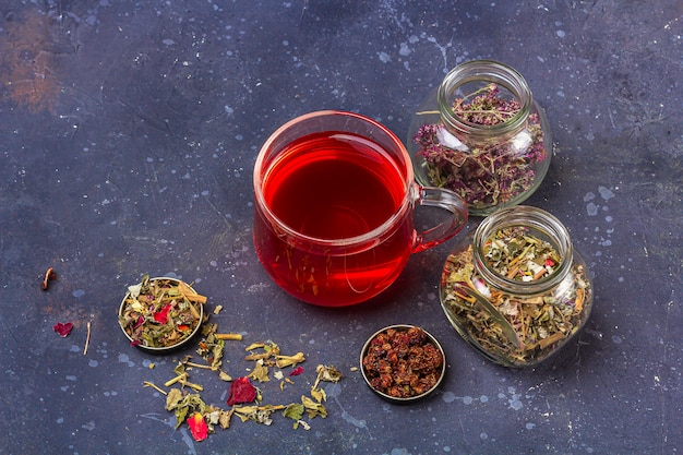 Фото Красный чай (ройбуш, гибискус, каркаде) в стеклянной чашке и банки сухих чайных листьев и лепестков на темном фоне. травяной, витаминный, детокс чай от простуды и гриппа
