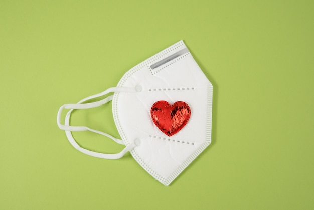 Red textile small heart lies on a white disposable medical mask, green surface