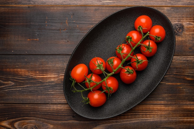 Red tomatoes on a green branch on a dark platter on a wooden background.