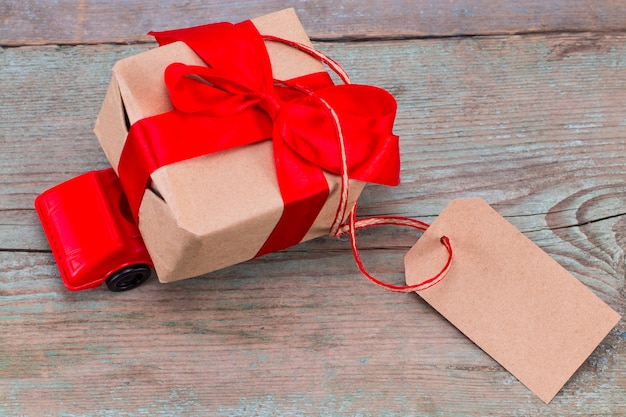 Red toy car delivering gifts box with tag with empty space for a text on wooden background.