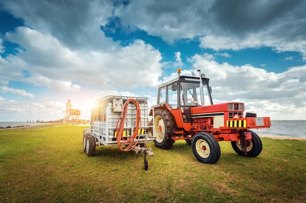 Red tractor with trailer on the grass field against lighthouse and blue cloudy sky in spring at sunset. Agricultural tractor. Agricultural machinery and machines in bright sunny day. Landscape