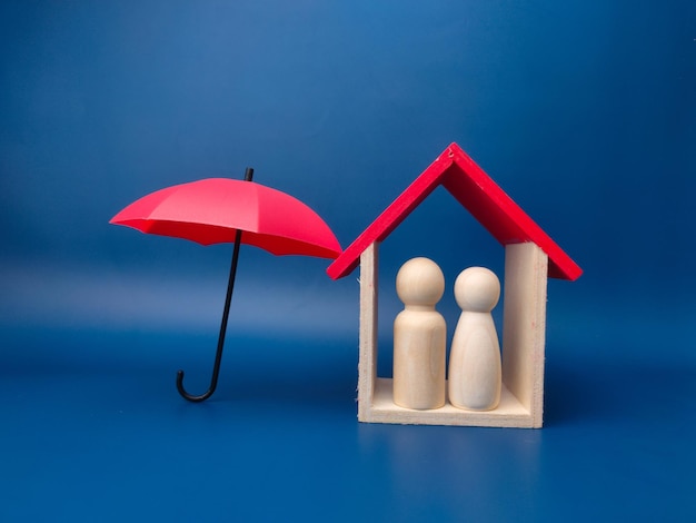 Red umbrella protect wooden house and peg doll on a blue\
background planning saving families preventing risks and crises\
health care and insurance concepts