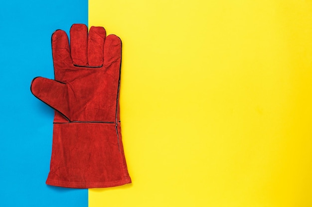 Red welder's glove from the left hand on a yellow and blue background Protective accessory for welding operations