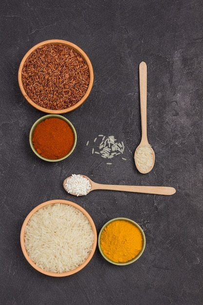 Red and white rice in bowls. Turmeric and ground red pepper. White rice in wooden spoon. Black surface. Flat lay