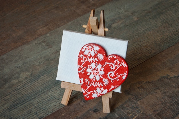 Красное деревянное сердце с нарисованным узором стоит на небольшом штативе с чистым белым холстом.