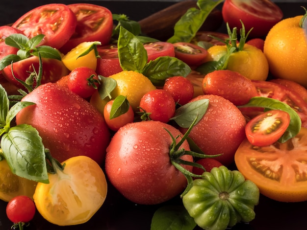 さまざまな種類の赤と黄色のトマトがバジルと一緒にテーブルの上にあり、それらのいくつかはカットされています。