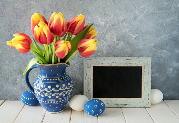 Red-yellow tulips in blue ceramic pitcher with Easter eggs and a blackboard, text space