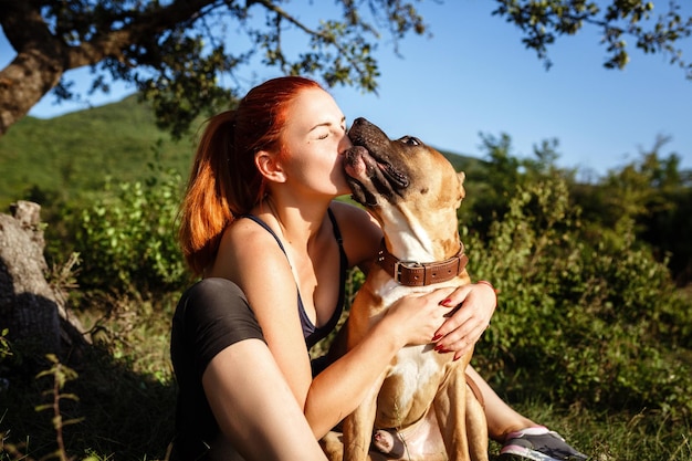 日当たりの良い公園で彼らの時間と休暇を楽しんでいるスポーツ服を着て犬を愛撫する赤毛の陽気な若い女性