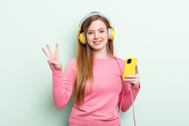 redhair woman smiling and looking friendly, showing number three. headphones and smartphone concept