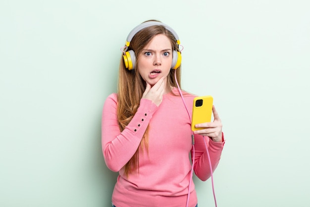 Donna dai capelli rossi con la bocca e gli occhi spalancati e la mano sul mento. concetto di cuffie e smartphone