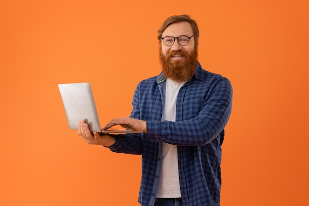 赤のひげのビジネスマンがオレンジ色の背景でラップトップのコンピューターで働いています