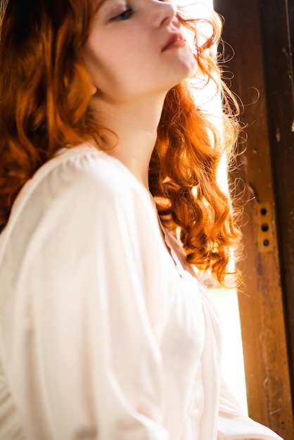 Redhaired beautiful girl near the old window in a white night dress in a French traditional house the theme of romance