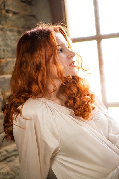 Redhaired beautiful girl near the old window in a white night dress in a French traditional house the theme of romance