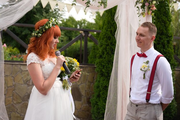 Photo redhaired bride with flowers reads from the phone into the microphone to groom in a white shirt