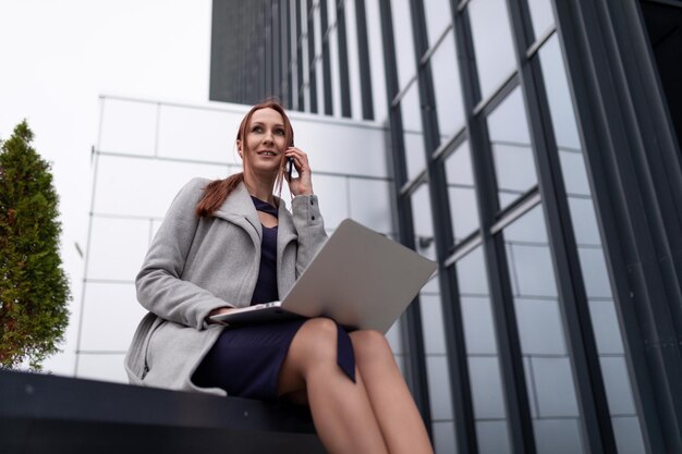 Redhaired businesswoman with a laptop talking on a mobile phone outside the office