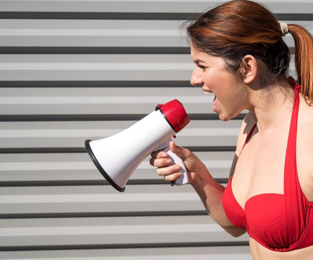 Redhaired Caucasian woman in a red bikini shouting into a megaphone against a background of a gray corrugated wall Girl in a swimsuit with a loudspeaker Summer vacation