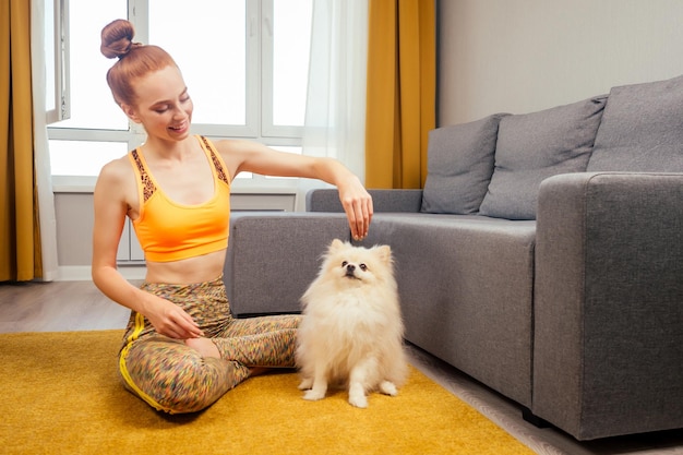 Redhaired ginger girl having fun with her cute fluffy spitz on the yellow carpet in a stylish spacious apartment.
