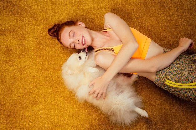 Redhaired ginger girl in a stylish top and leggings having fun with her cute fluffy spitz on the yellow carpet in a stylish spacious apartment