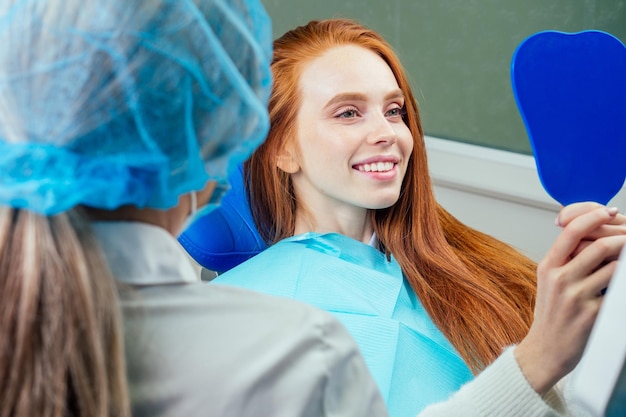 Redhaired ginger lady checking teeth in mirror at dentist office