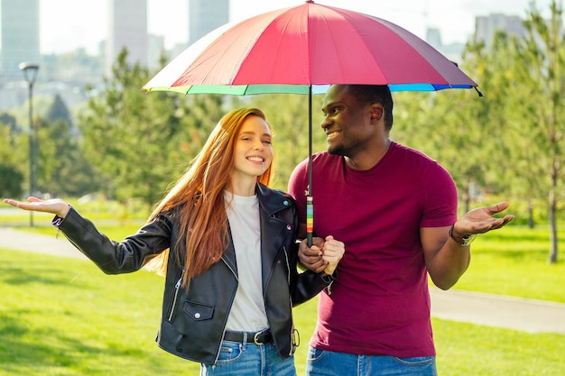 Redhaired ginger woman and african american man standing close to each other under umbrella in summer park