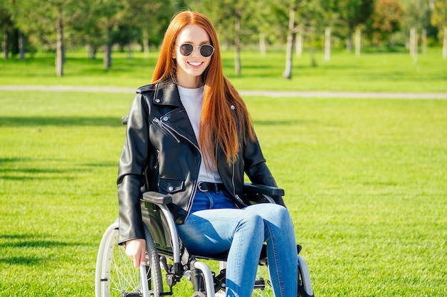 写真 幸せな赤毛の生姜の女性、彼女は車椅子に座って、サングラスと暖かい革のジャケットを夏の公園で着ています。