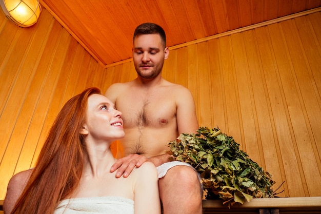 Redhaired ginger woman and her boyfriend steaming in the bathhouse with russian bunch broomcouple in love enjoying massage the sauna together at the spa