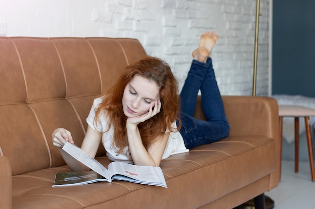Рыжая девушка лежит на диване и читает дома журнал
