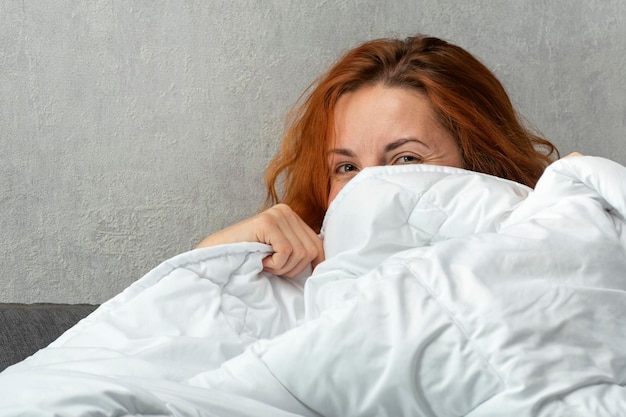 Redhaired girl shyly peeks out from under the covers Portrait of a beautiful young women