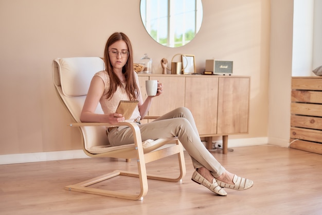 redhaired girl sits in an armchair and holds a cup and looks into sketchpad in cozy interior
