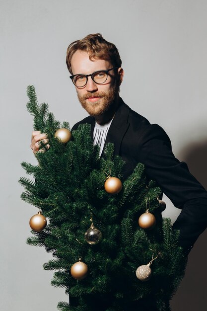 Redhaired handsome man in a black jacket and glasses dances with a Christmas tree in his hands New Year party concept