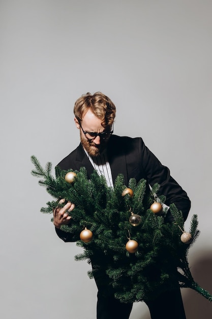 Redhaired handsome man in a black jacket and glasses dances with a Christmas tree in his hands New Year party concept