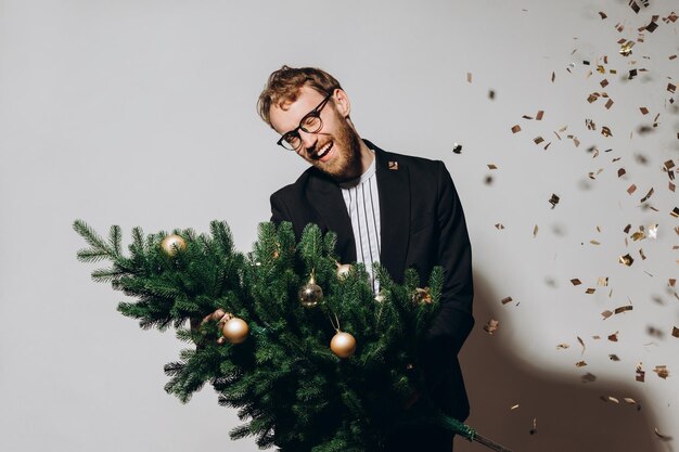 Redhaired handsome man in a black jacket and glasses dances with a Christmas tree in his hands New Year party concept