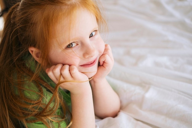 A redhaired little girl in a green Tshirt lies on a white bed at home and smiles